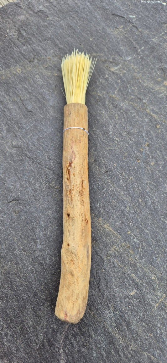 A pastry brush made from pale yellow tampico plant fibre, white waxed linen and a driftwood handle lies on a slate surfact